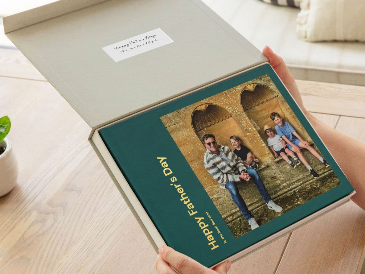 A person opens a presentation box revealing a photo book titled "Happy Father's Day" with a cover photo of three people sitting on stone steps.