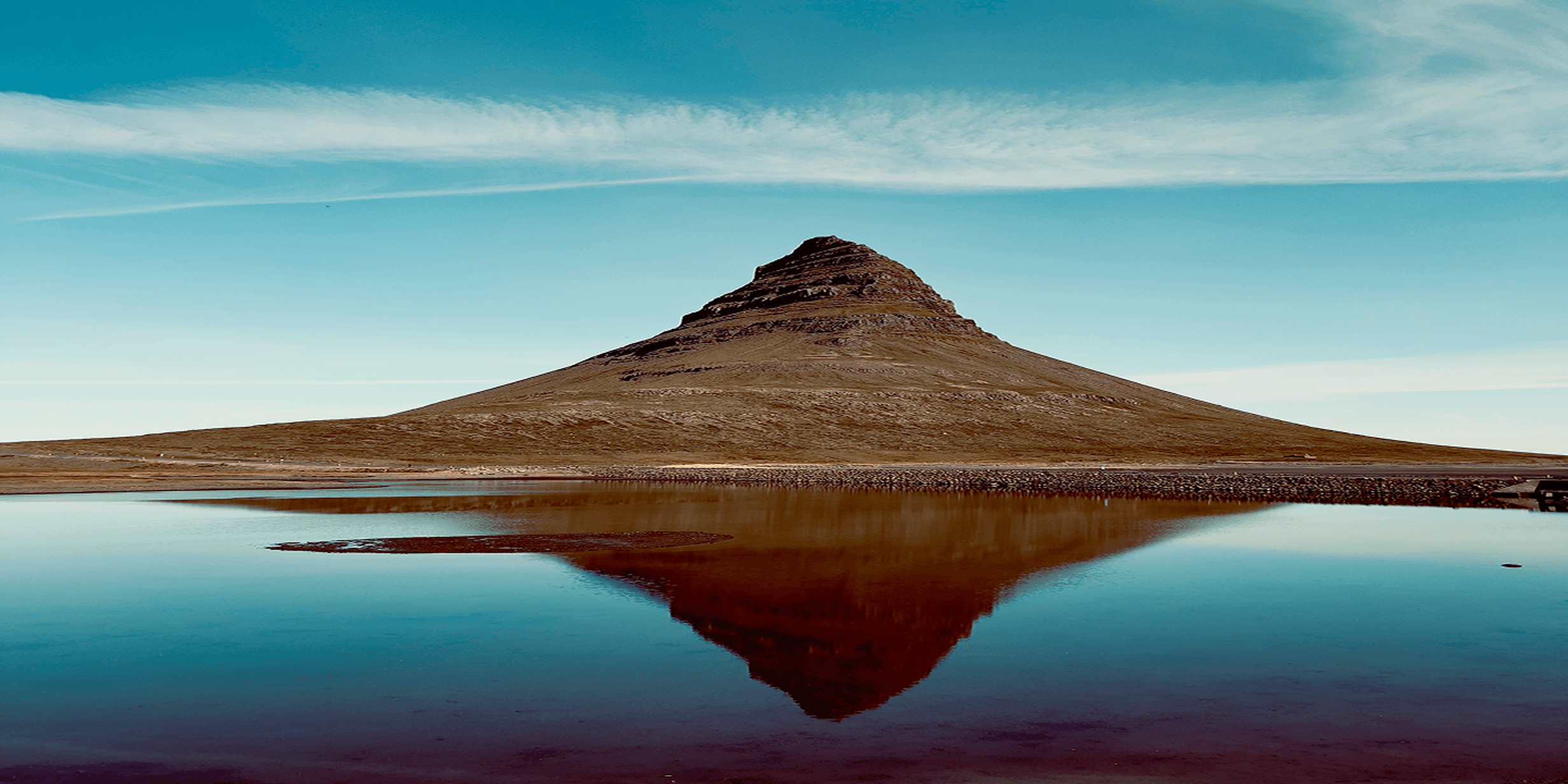 A conical mountain reflected in a calm lake under a clear blue sky, with a thin layer of clouds stretching horizontally.