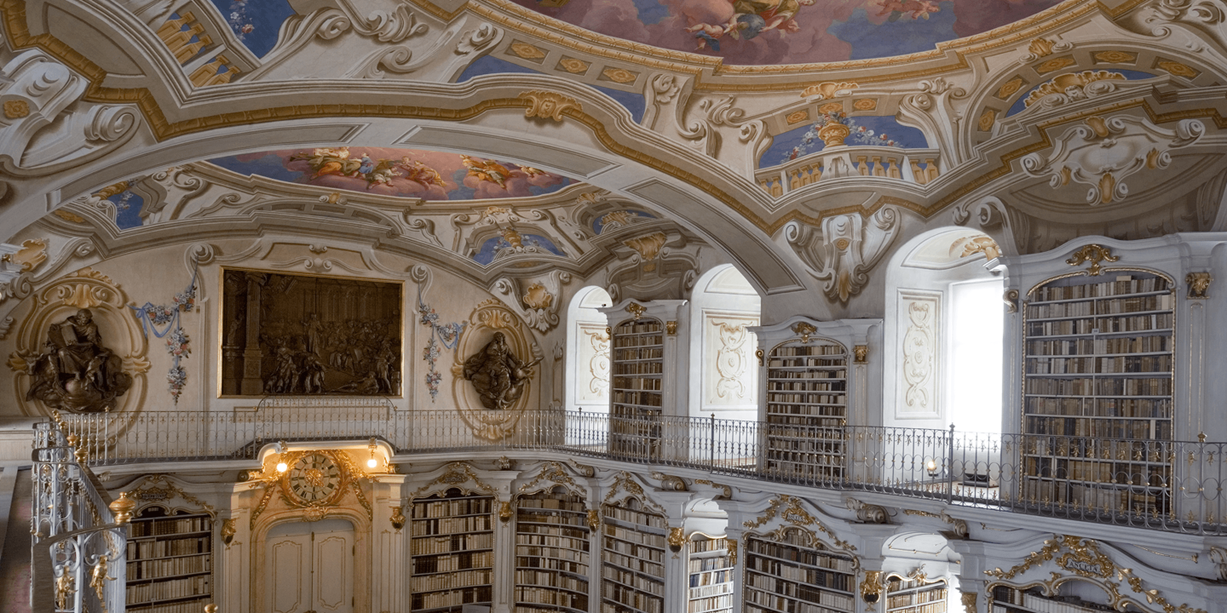 Ornate library interior with arched ceiling, elaborate frescoes, tall bookshelves, and decorative sculptures. Bright natural light illuminates the space.