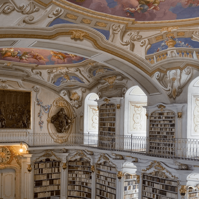Ornate library interior with arched ceiling, elaborate frescoes, tall bookshelves, and decorative sculptures. Bright natural light illuminates the space.