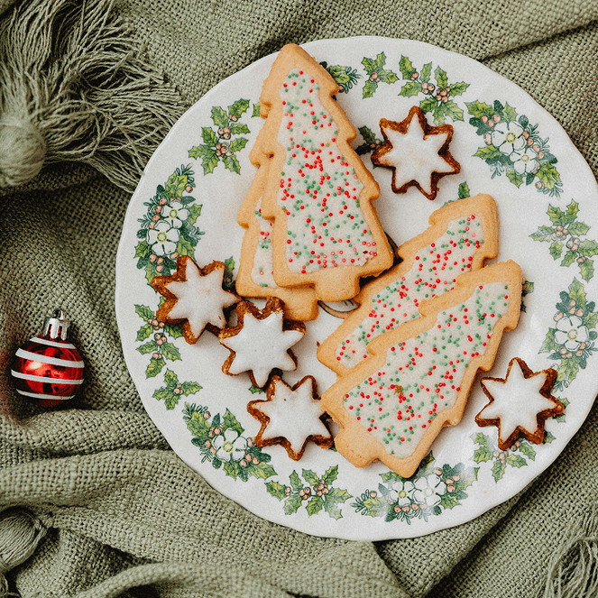 Plate of Christmas-themed cookies on a green cloth, surrounded by red ornaments and a nutcracker figurine.