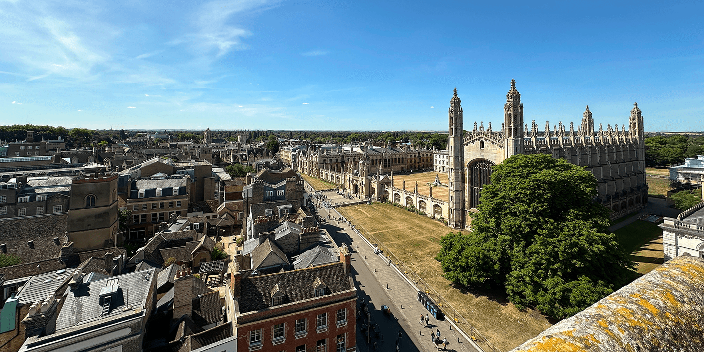 Aerial view of a historic city with a large Gothic-style building, tree-lined streets, and a clear blue sky.
