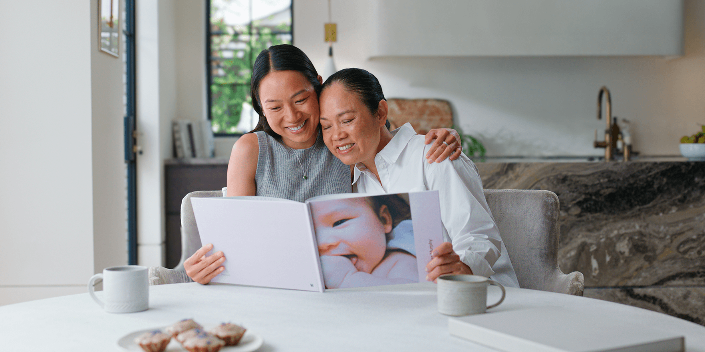Two women sitting at a table, smiling and looking at a photo album in a bright kitchen