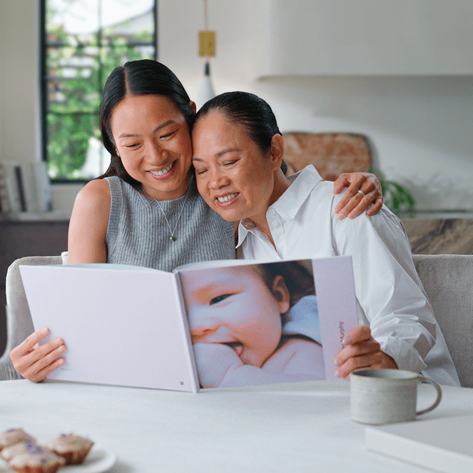 Two women sitting at a table, smiling and looking at a photo album in a bright kitchen