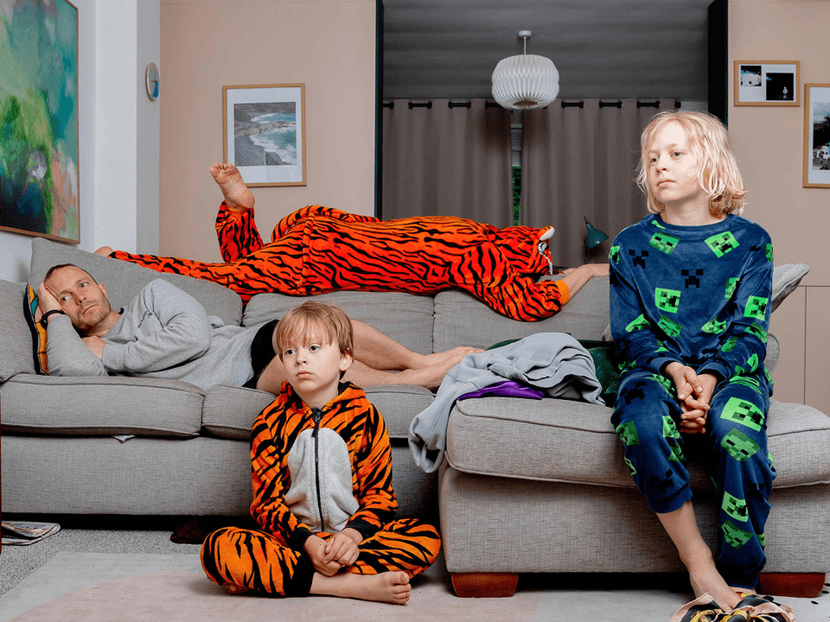 Three people in colorful pajamas relax in a living room; one lies on a couch, while two children sit on the floor and couch.