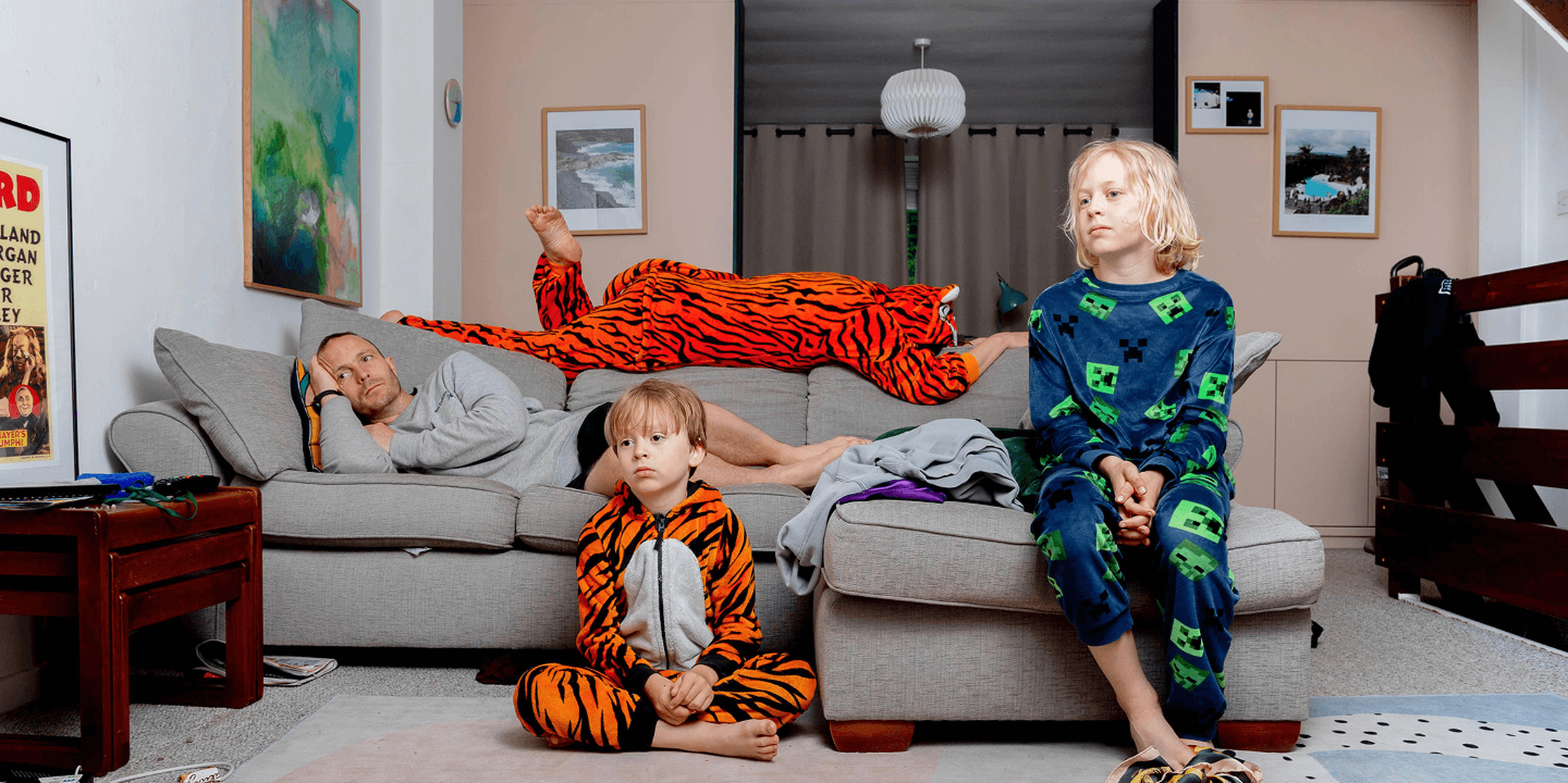 Three people in colorful pajamas relax in a living room; one lies on a couch, while two children sit on the floor and couch.