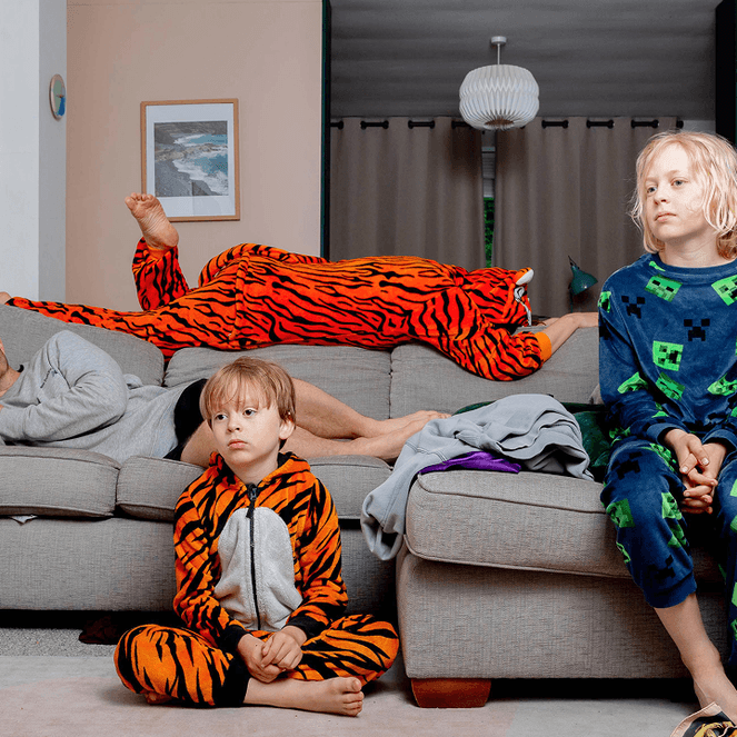 Three people in colorful pajamas relax in a living room; one lies on a couch, while two children sit on the floor and couch.