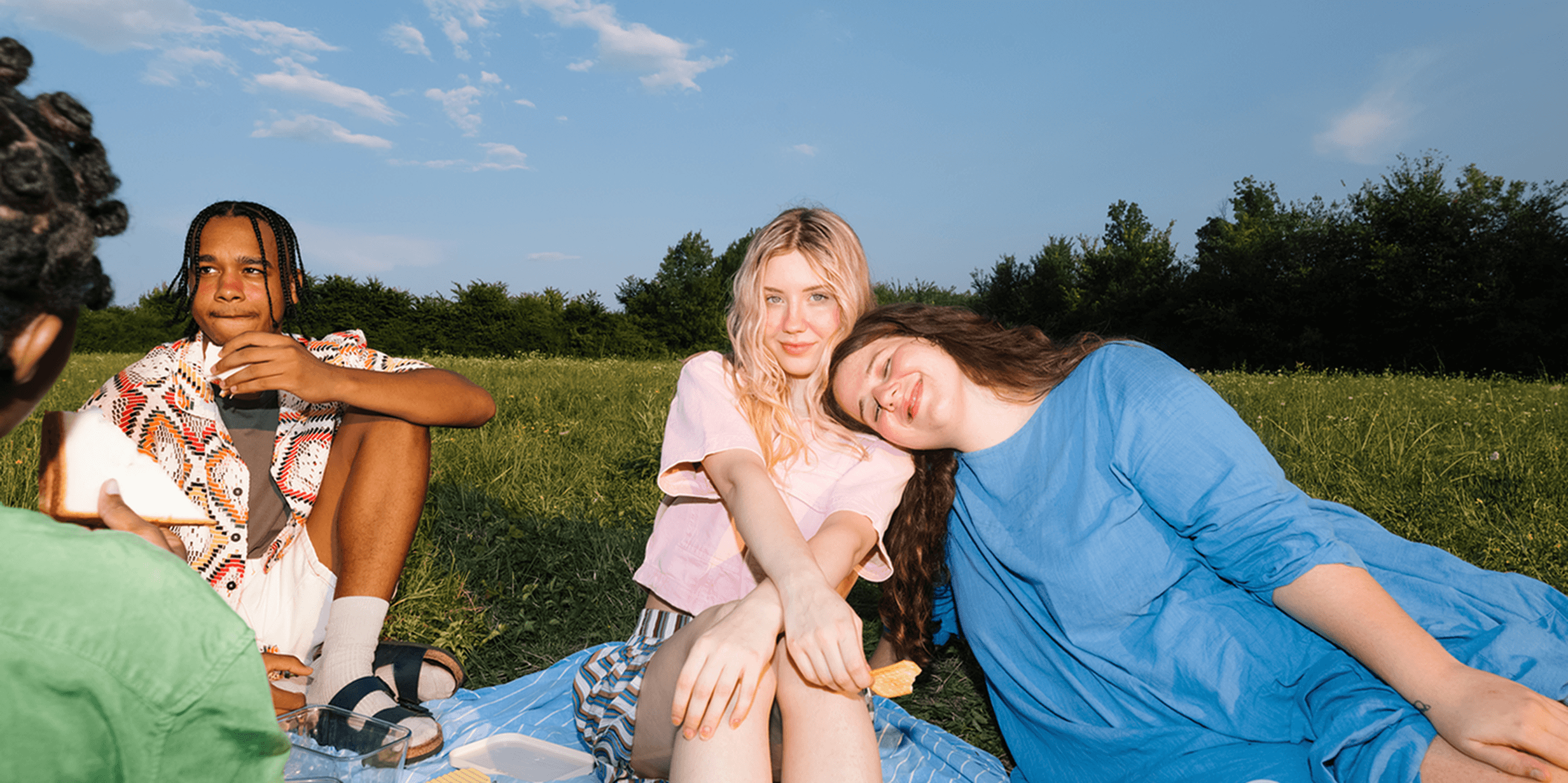 Three people enjoying a sunny picnic on a grassy field, with one person resting their head on another's shoulder, surrounded by trees.