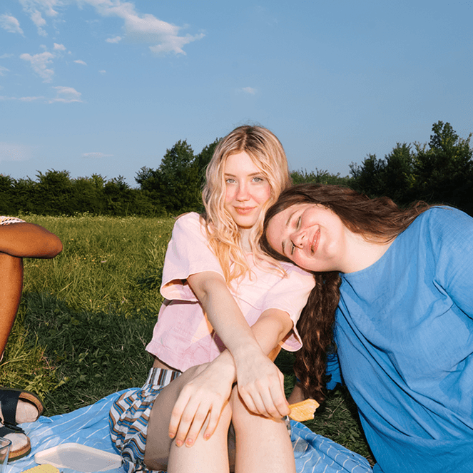 Three people enjoying a sunny picnic on a grassy field, with one person resting their head on another's shoulder, surrounded by trees.