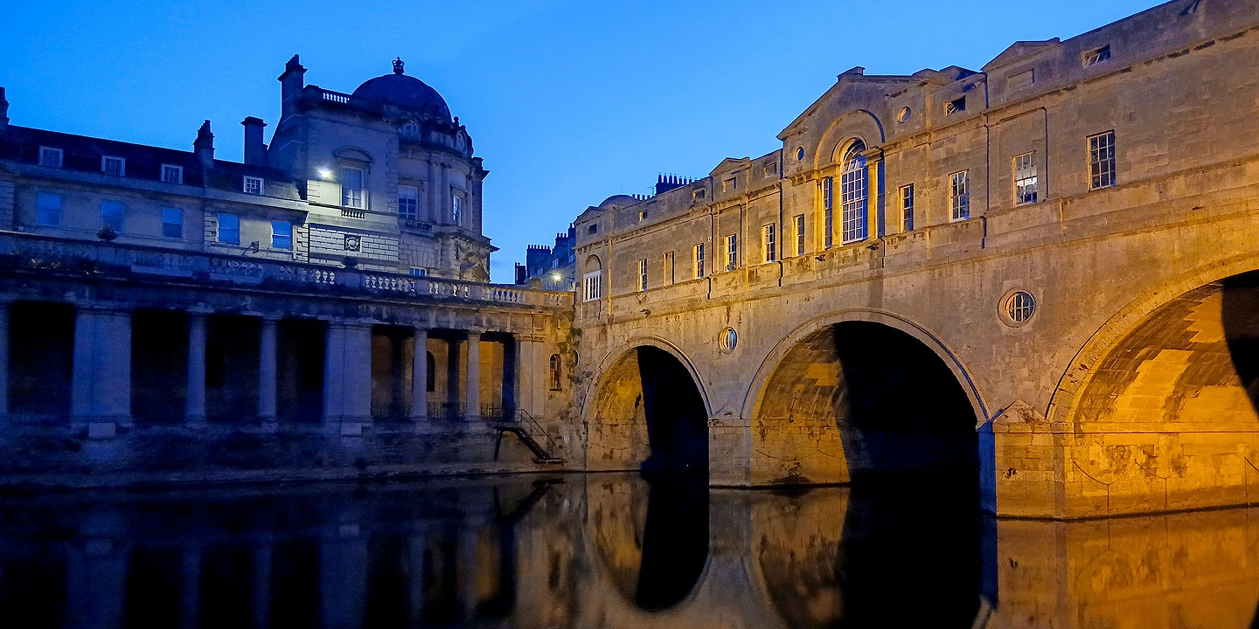 Illuminated historic stone bridge and buildings reflected in calm river water at twilight, under a clear blue sky.