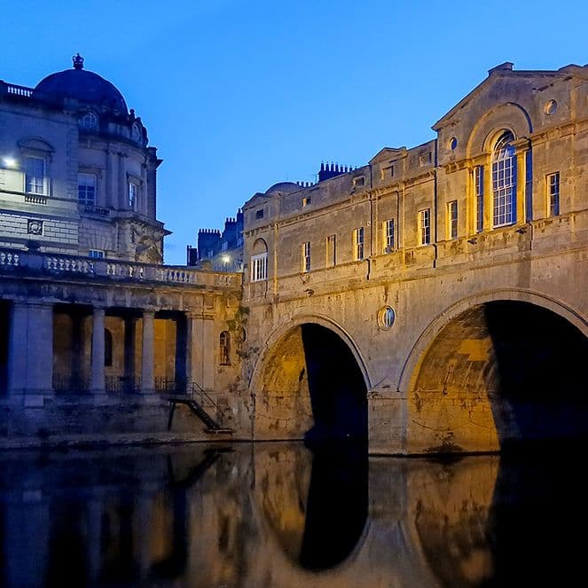 Illuminated historic stone bridge and buildings reflected in calm river water at twilight, under a clear blue sky.