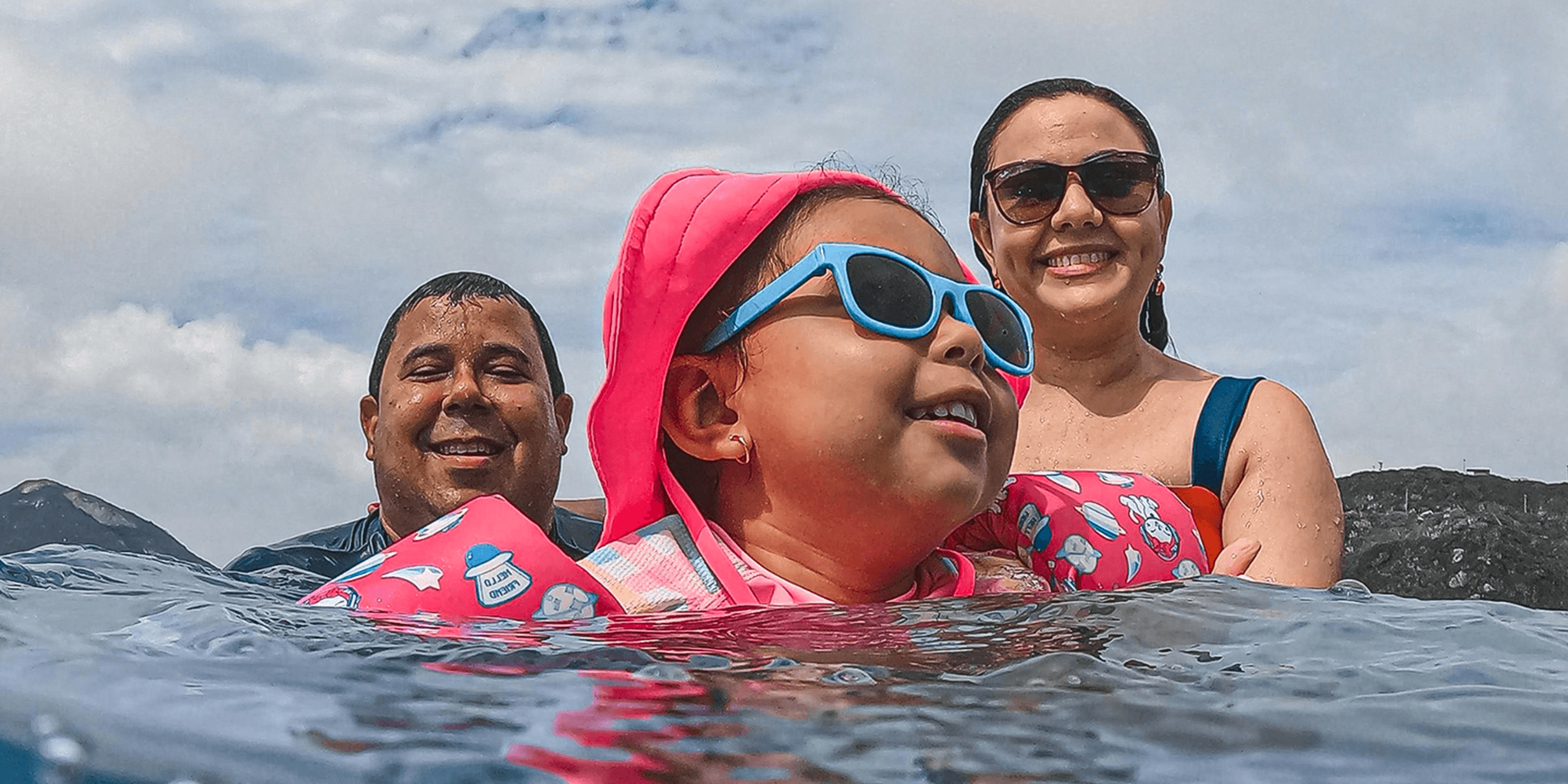A family of three enjoys swimming in the sea. The child wears pink gear and blue sunglasses, with the parents smiling in the background.