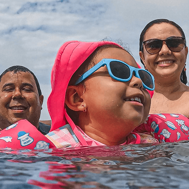 A family of three enjoys swimming in the sea. The child wears pink gear and blue sunglasses, with the parents smiling in the background.