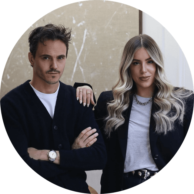 A man and woman stand together indoors, both wearing dark cardigans. The woman has long, wavy hair, and the man has short, styled hair.