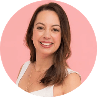 Smiling woman with long brown hair wearing a white top and necklace, set against a pink background.