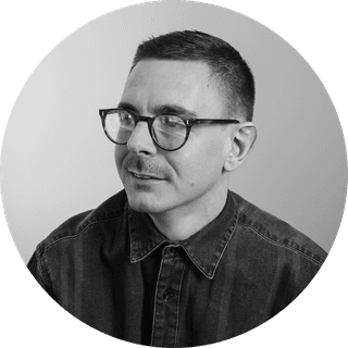 Man with short hair and glasses, wearing a dark button-up shirt, looking to the side against a plain background.