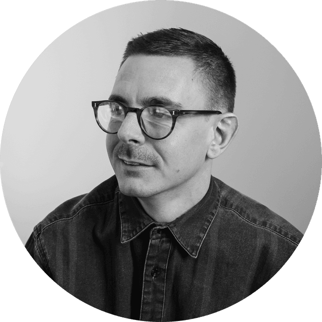 Man with short hair and glasses, wearing a dark button-up shirt, looking to the side against a plain background.