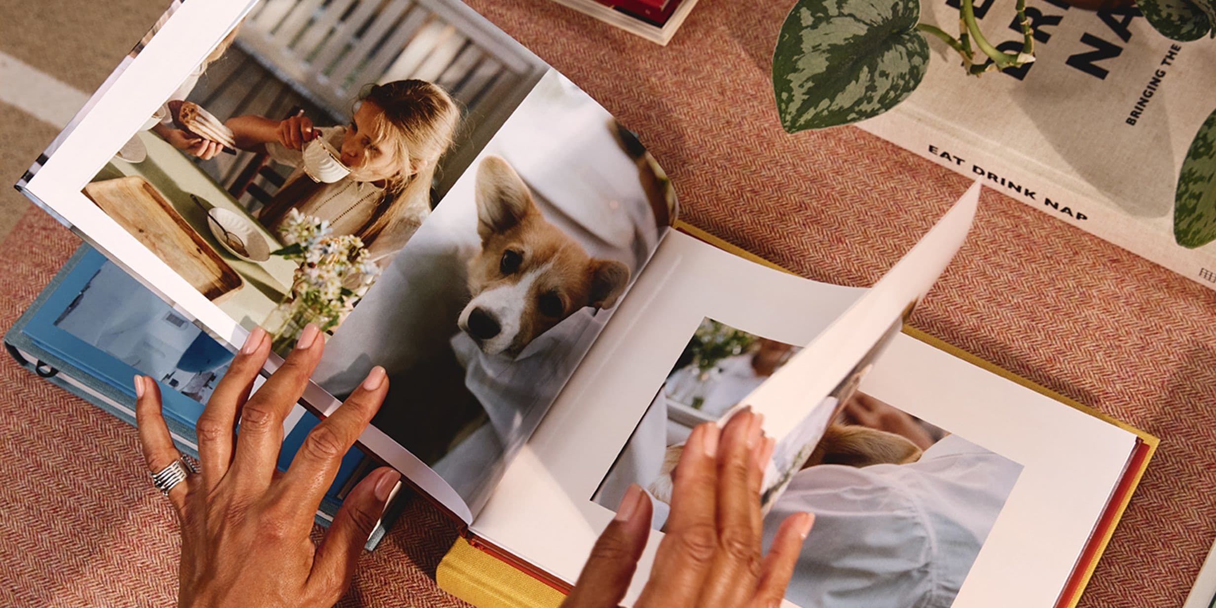 Hands flipping through a photo book showing images of a woman drinking coffee and a dog. Nearby, a book titled "Eat Drink Nap."