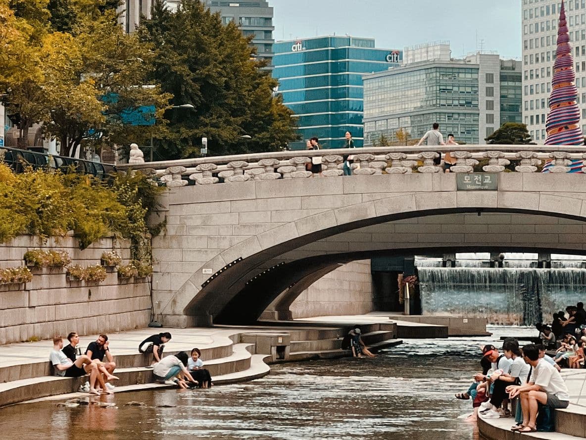 People relax by a riverside under a stone bridge in an urban park, surrounded by trees and modern buildings.