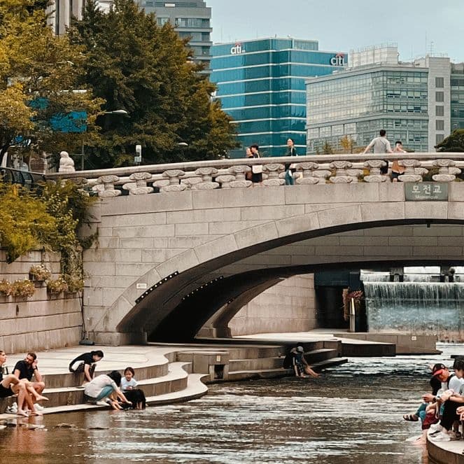 People relax by a riverside under a stone bridge in an urban park, surrounded by trees and modern buildings.
