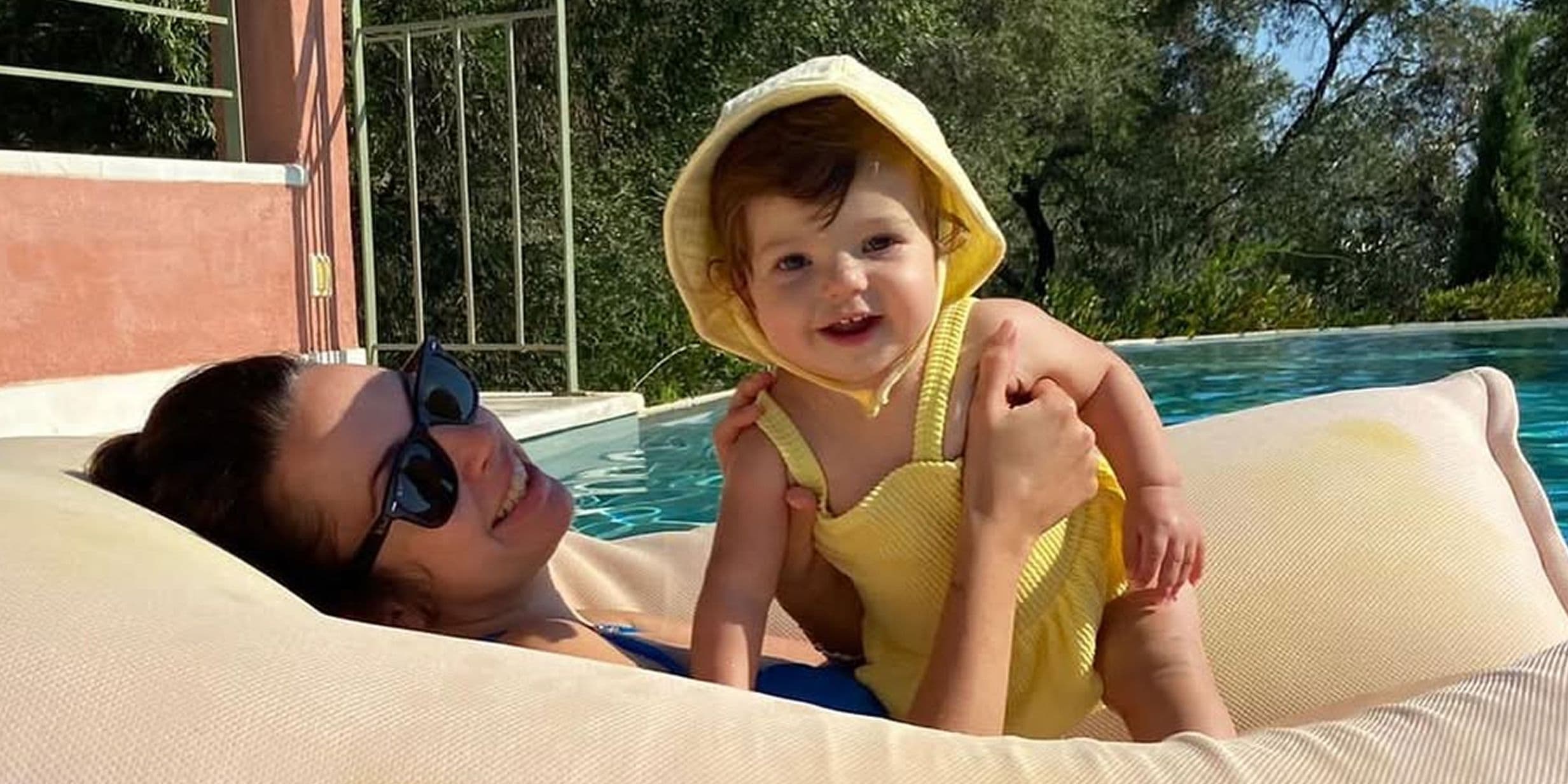 Woman lounging on a pool float, holding a smiling baby in a yellow outfit with a hood. Trees and a building are in the background.