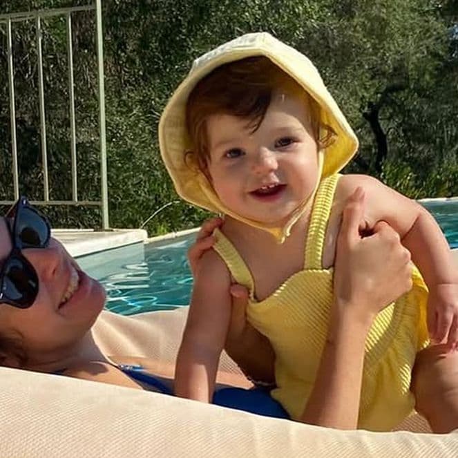 Woman lounging on a pool float, holding a smiling baby in a yellow outfit with a hood. Trees and a building are in the background.