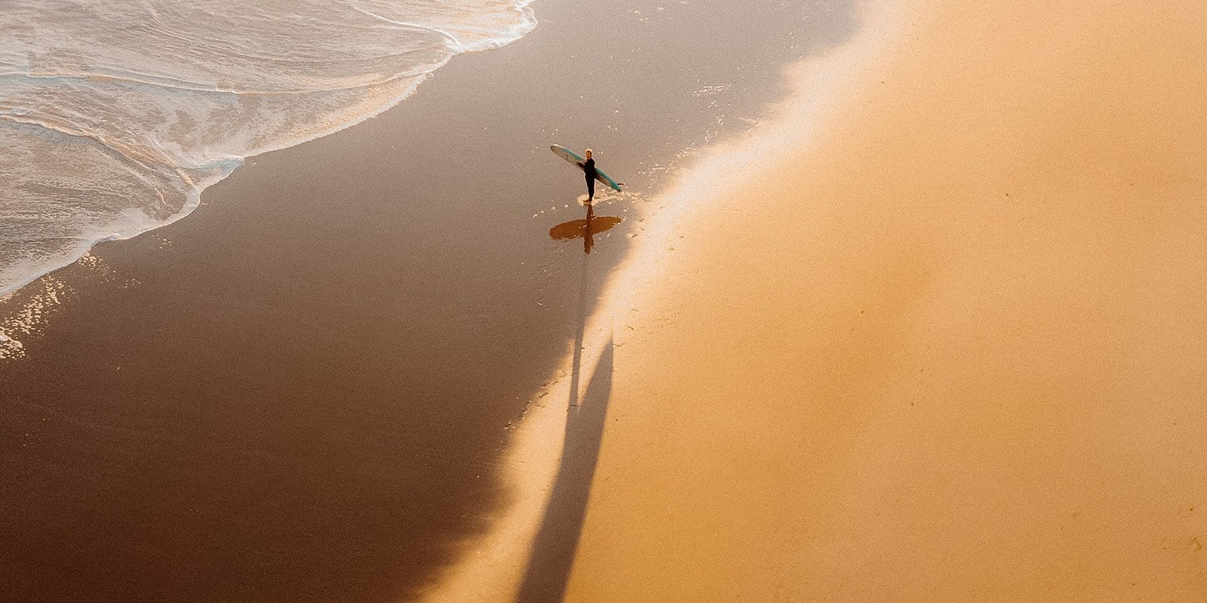 An aerial photogrpah of a surfer approaching the water from a sandy beach, shot from a drone