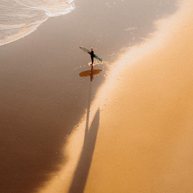 An aerial photogrpah of a surfer approaching the water from a sandy beach, shot from a drone
