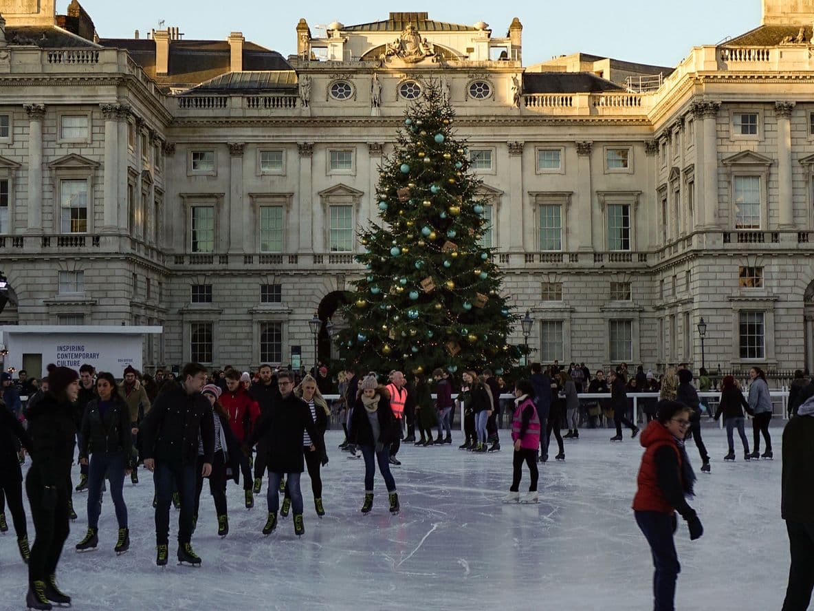 People ice skating in front of a large Christmas tree at an outdoor rink, surrounded by Somerset House's historic buildings and festive lights.