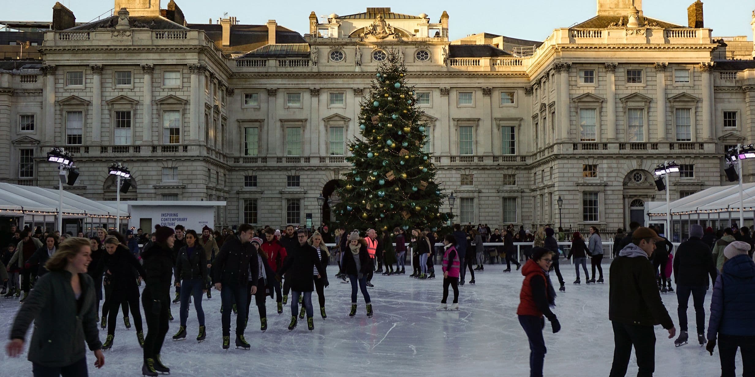 People ice skating in front of a large Christmas tree at an outdoor rink, surrounded by Somerset House's historic buildings and festive lights.