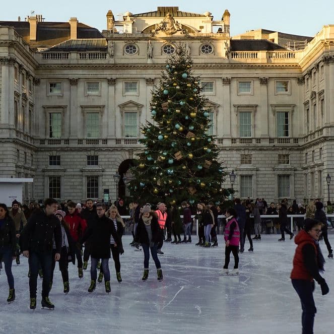 People ice skating in front of a large Christmas tree at an outdoor rink, surrounded by Somerset House's historic buildings and festive lights.