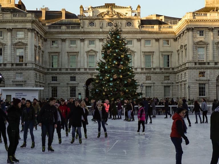 People ice skating in front of a large Christmas tree at an outdoor rink, surrounded by Somerset House's historic buildings and festive lights.