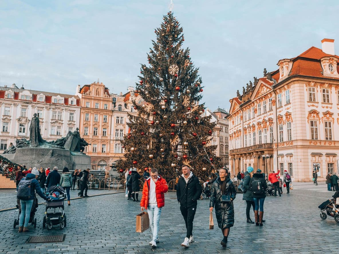 People walk around a large decorated Christmas tree in a busy European square surrounded by historic buildings.