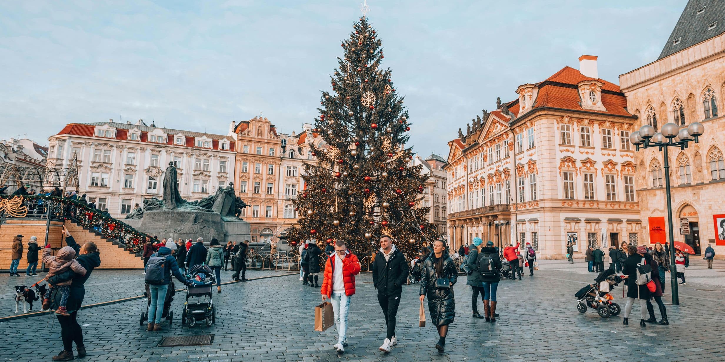 People walk around a large decorated Christmas tree in a busy European square surrounded by historic buildings.