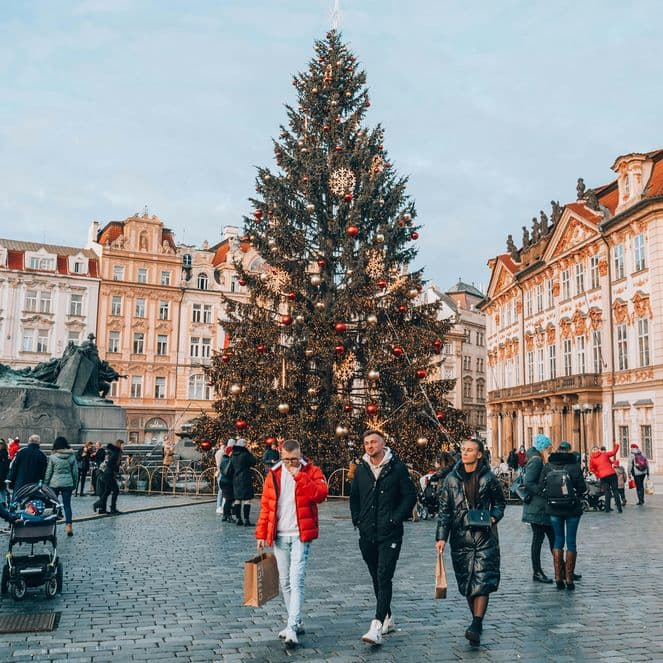 People walk around a large decorated Christmas tree in a busy European square surrounded by historic buildings.