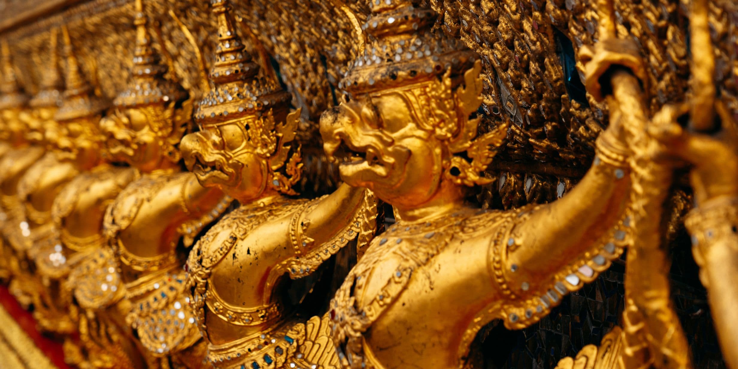 Close-up of gilded mythic guardian figures with ornate crowns and jewelry carved in a row on a temple facade.