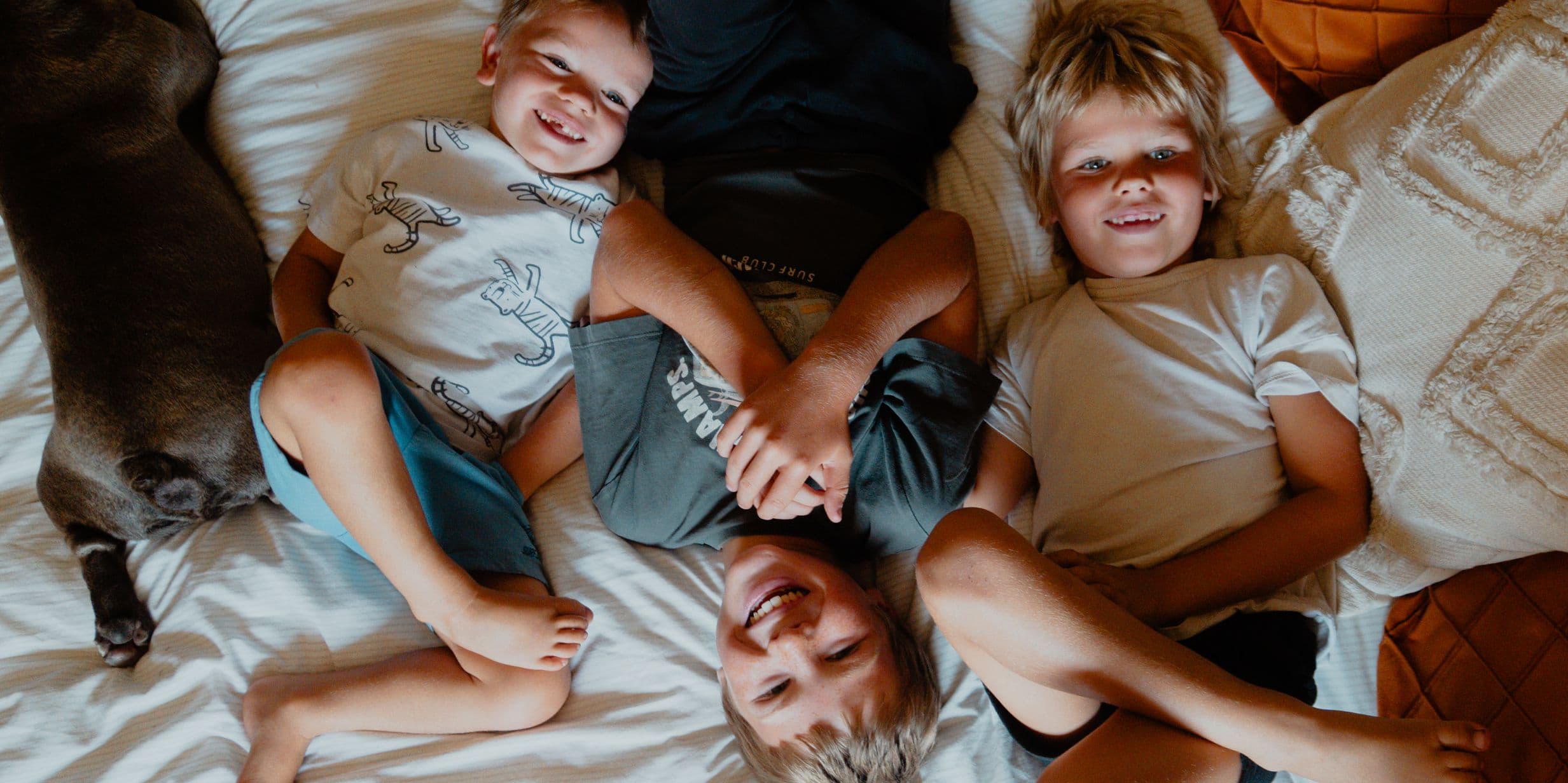 Three children lying on a bed, smiling and playing together, with a dog resting nearby.