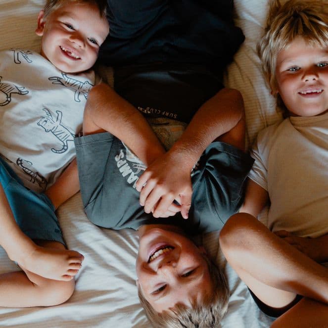 Three children lying on a bed, smiling and playing together, with a dog resting nearby.