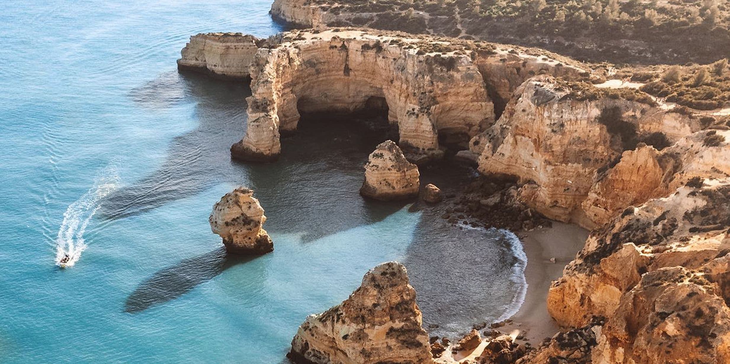 Aerial view of rocky cliffs and clear blue waters along a coastline, with a small boat leaving a wake near the shore.