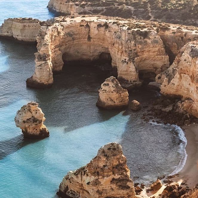 Aerial view of rocky cliffs and clear blue waters along a coastline, with a small boat leaving a wake near the shore.