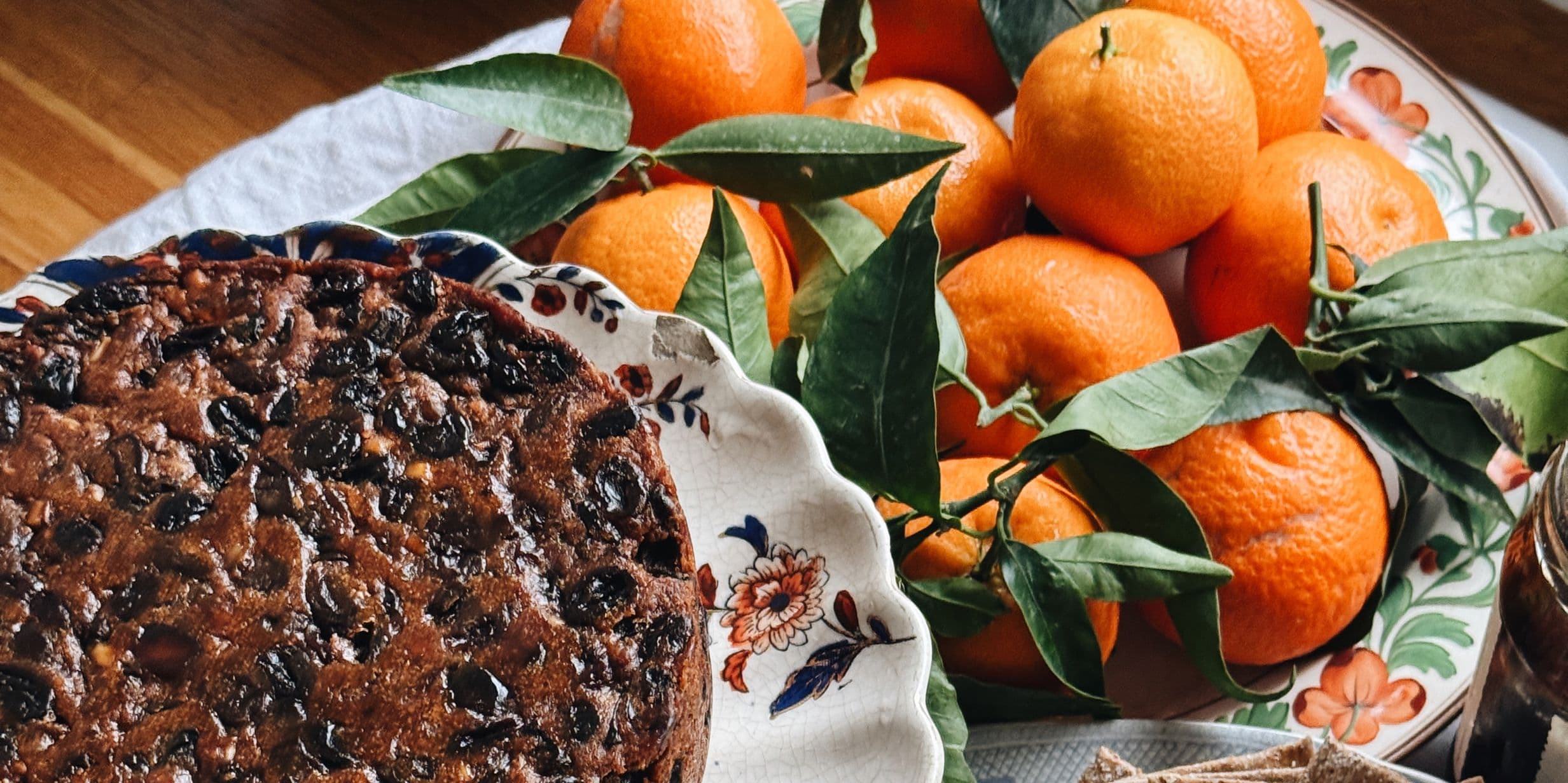 Fruitcake on a floral plate next to a bowl of fresh oranges with green leaves on a wooden table.