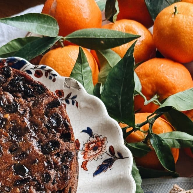 Fruitcake on a floral plate next to a bowl of fresh oranges with green leaves on a wooden table.