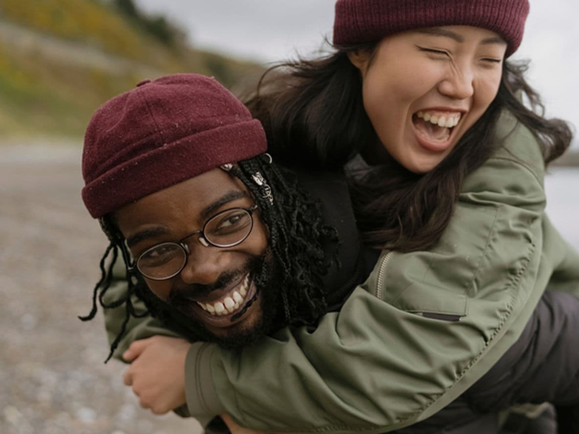 A joyful couple at the beach, one giving the other a piggyback ride. Both are wearing beanies and jackets, smiling and laughing.