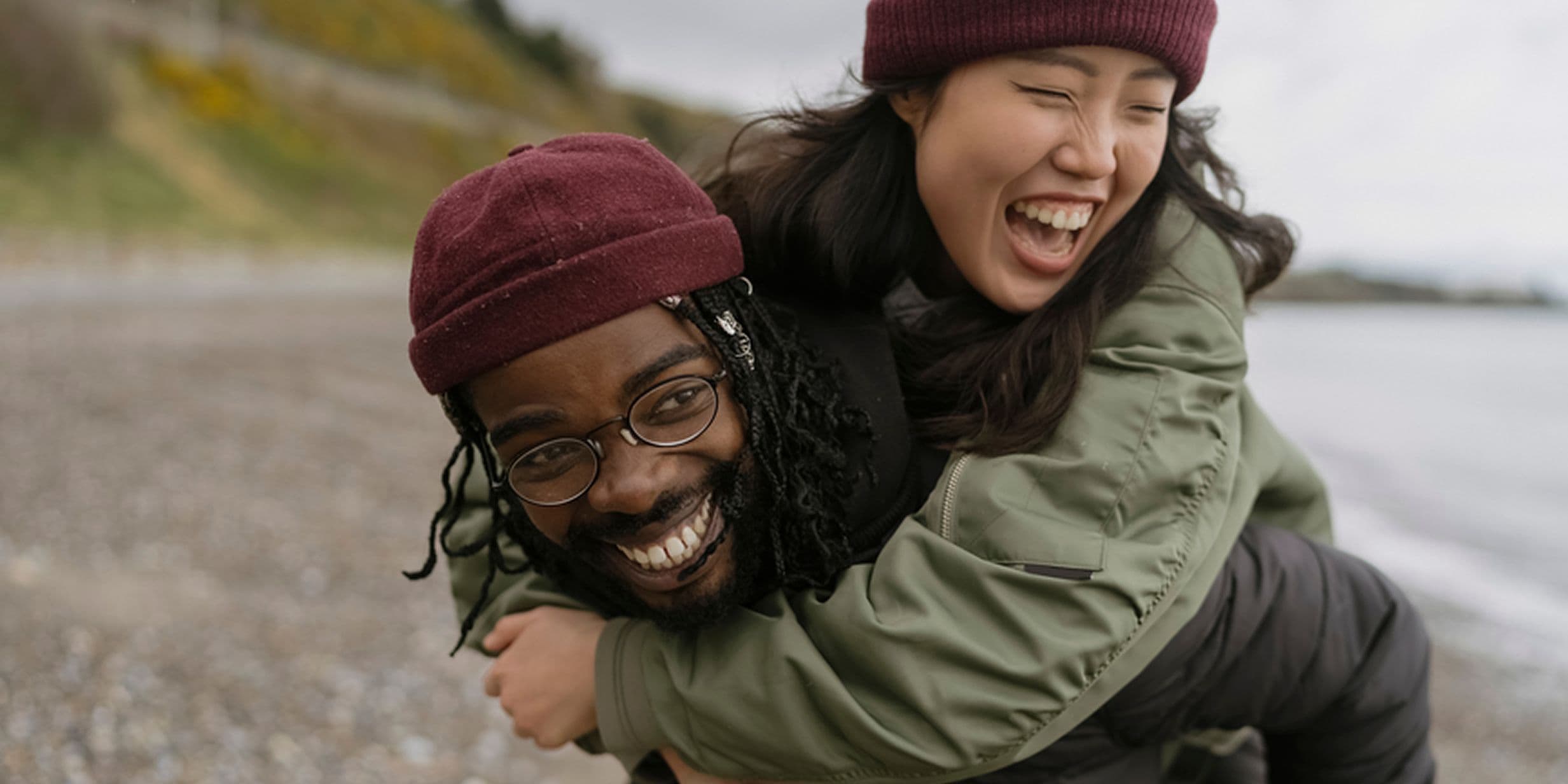 A joyful couple at the beach, one giving the other a piggyback ride. Both are wearing beanies and jackets, smiling and laughing.