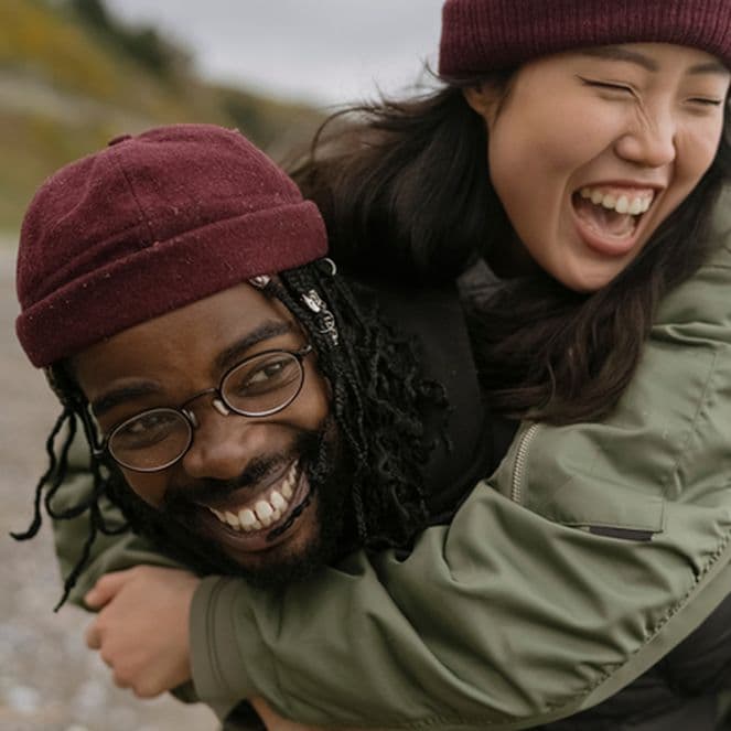 A joyful couple at the beach, one giving the other a piggyback ride. Both are wearing beanies and jackets, smiling and laughing.