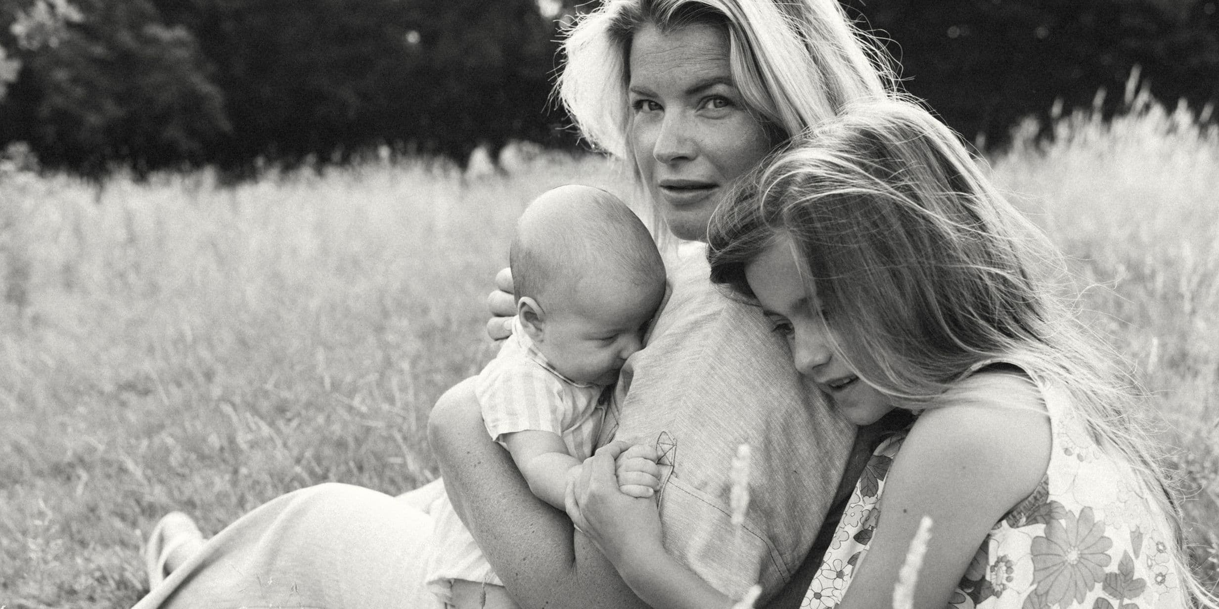 A woman sits in a field, holding a baby and embracing a young girl. The scene is serene and intimate, with tall grass in the background.