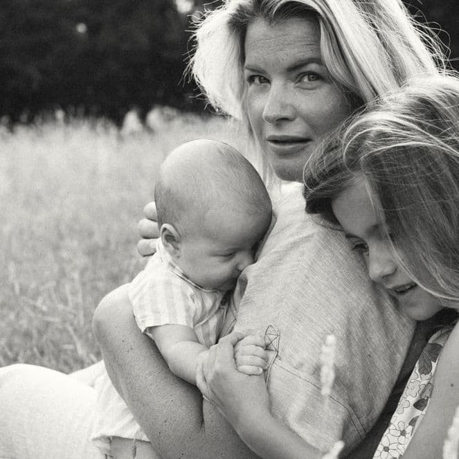 A woman sits in a field, holding a baby and embracing a young girl. The scene is serene and intimate, with tall grass in the background.