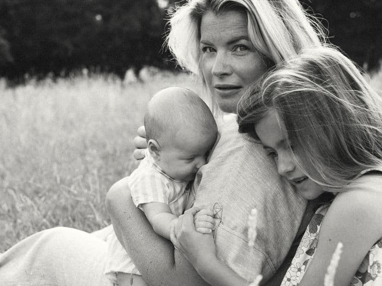A woman sits in a field, holding a baby and embracing a young girl. The scene is serene and intimate, with tall grass in the background.