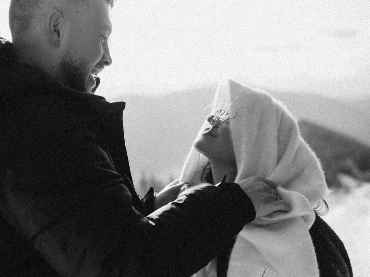 A man and woman smiling at each other on a snowy mountain trail. The woman is wrapped in a blanket. Black and white photo.