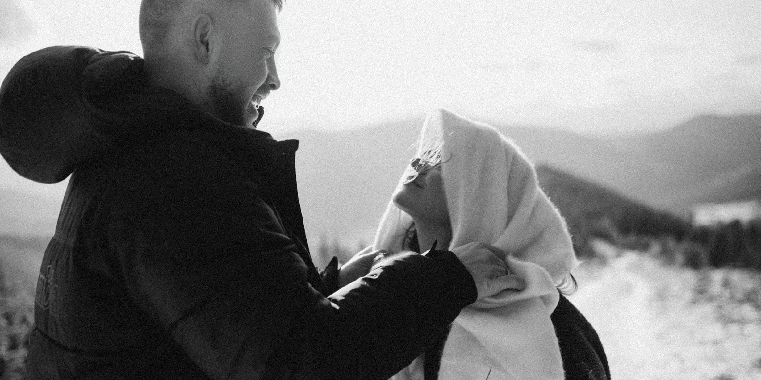 A man and woman smiling at each other on a snowy mountain trail. The woman is wrapped in a blanket. Black and white photo.